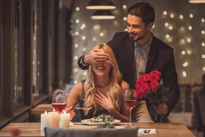 handsome elegant man is holding roses and covering his girlfriend's eyes while making a surprise in restaurant, both are smiling