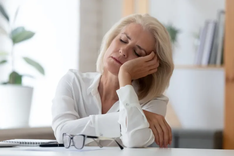 exhausted aged woman worker sit at office desk fall asleep distracted from work, tired senior businesswoman feel fatigue sleeping at workplace taking break dreaming or visualizing