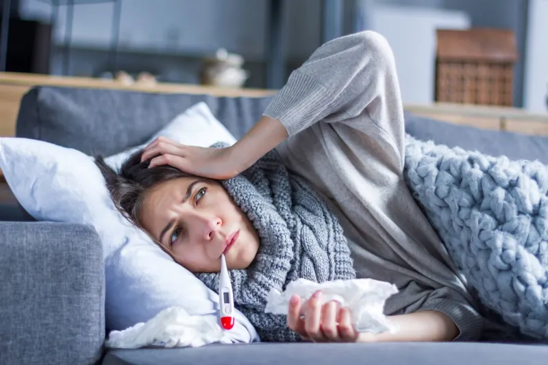 sick exhausted girl in scarf is lying in bed wrapped in blanket young woman with fever and headache is measuring temperature with thermometer, treated at home winter cold and flu concept