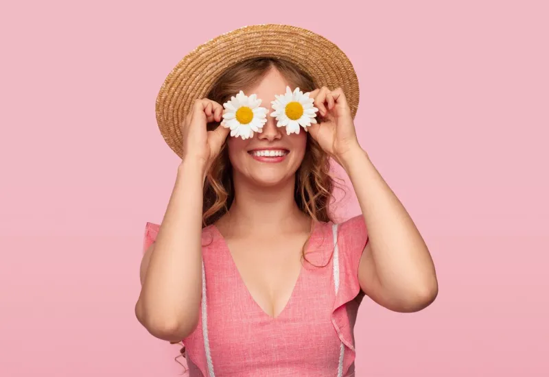 happy young female keeping fresh daisies near eyes and smiling while resting during summer vacation against pink background