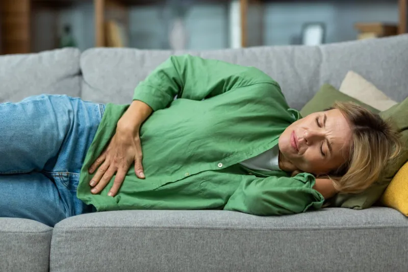 mature woman alone at home sick, holding hands to side of stomach having severe spasm lying on sofa in living room