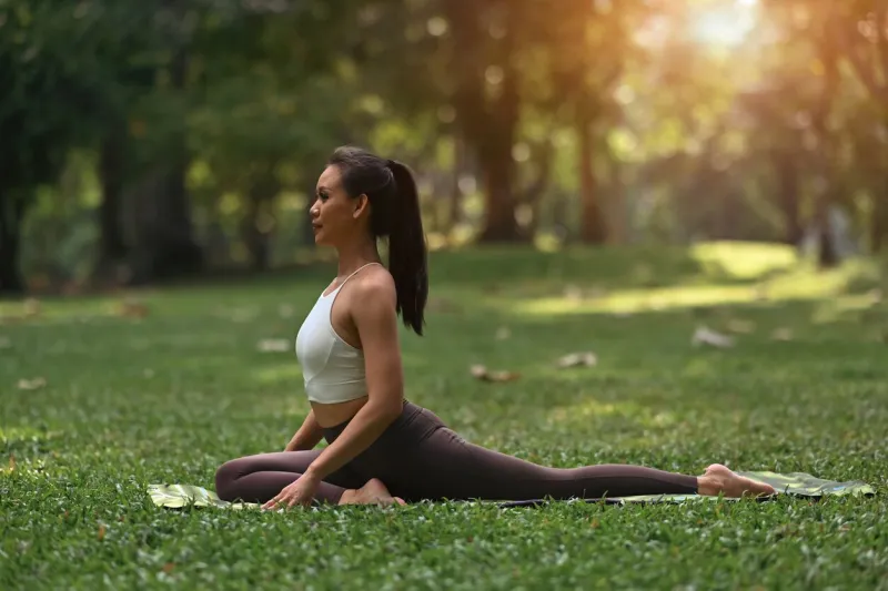 an asian woman demonstrates the pigeon pose (eka pada rajakapotasana), seated gracefully with one leg extended back, surrounded by vibrant greenery and soft, golden sunlight