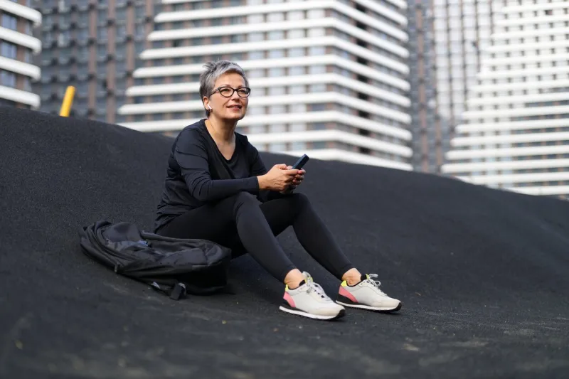 relaxed senior woman in sportswear with a smartphone in her hands is resting on the sports ground
