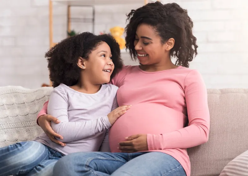 happy pregnant mother and her cute afro daughter hugging at home, sitting on couch and bonding together, little girl tenderly touching mom's belly