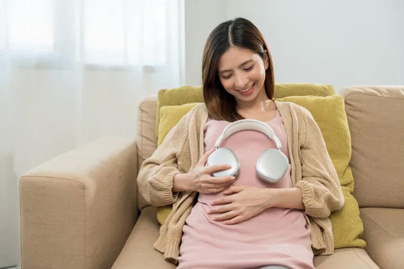 happy pregnant young woman holding headphone on her womb at home, relaxed young asian mother let her fetal listening to music, family planning and pregnancy with insurance for child health
