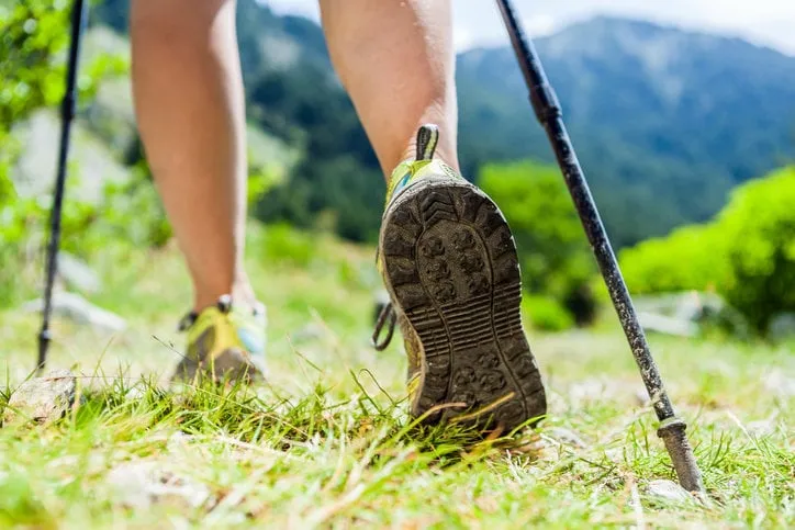 woman hiking in mountains, adventure and exercising nordic walking in sunny summer nature outdoors legs and sport shoes walk on grass