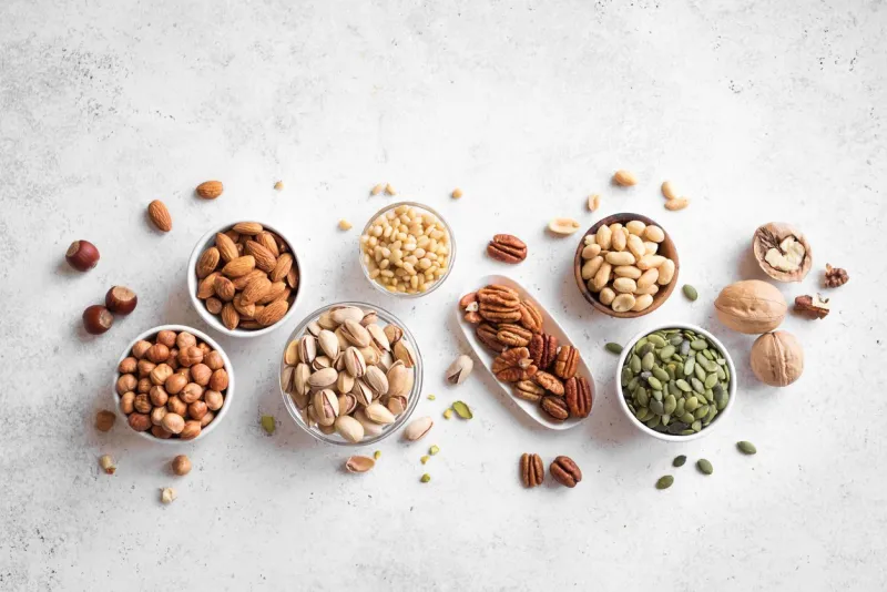 various nuts in bowls on white background, top view, copy space nuts assortment - pecans, hazelnuts, walnuts, pistachios, almonds, pine nuts, peanuts, pumpkin seeds