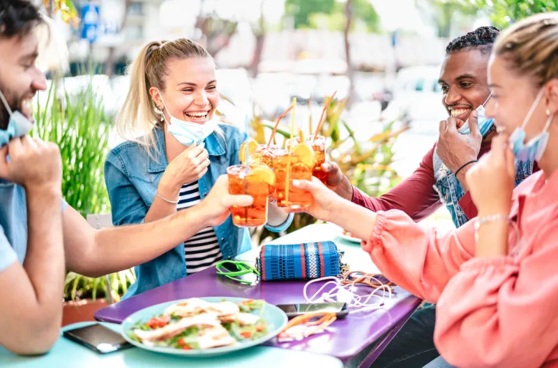 friends drinking spritz at cocktail bar with face masks - new normal friendship concept with happy people having fun together toasting drinks at restaurant - bright filter with focus on left woman