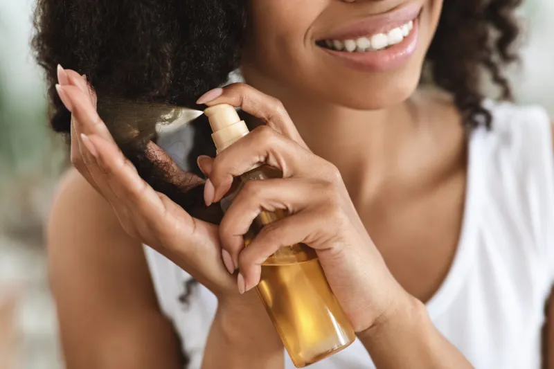 split ends repair treatment smiling african woman applying essential oil spray on her curly brown hair at home, cropped image, closeup