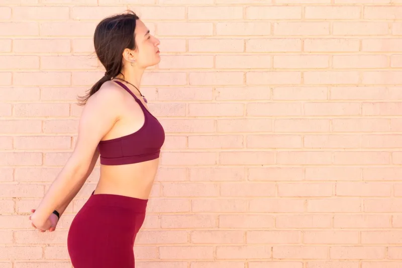 a young caucasian woman dressed in a top and leggings is doing arm, chest and back stretches next to a brick wall