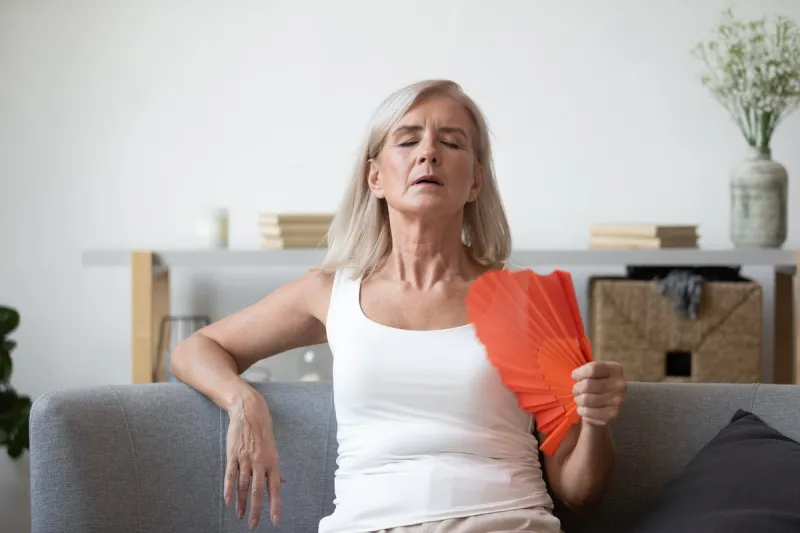 unwell elderly female retiree sit on couch feel dehydrated tired of heat use hand waver to cool down, overheated stressed elderly woman suffer from heatstroke in living room with no air conditioner