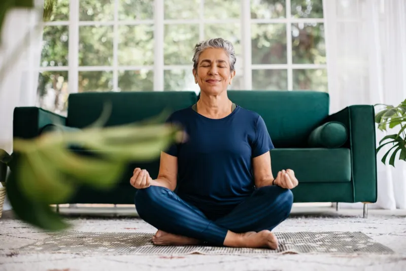 senior woman meditating in lotus position at home, sitting on the floor in fitness clothing mature woman doing a breathing workout to achieve relaxation, peace and mindfulness