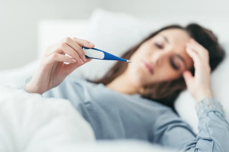 woman with flu virus lying in bed, she is measuring her temperature with a thermometer and touching her forehead