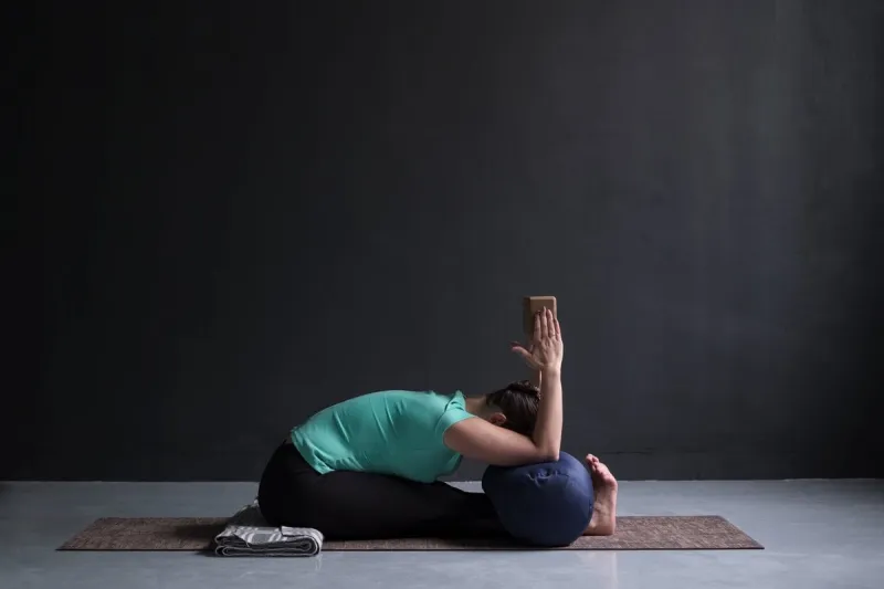 young woman practicing yoga, doing paschimottanasana exercise, seated forward bend pose, working out, using brick or block and bolster indoor full length photo