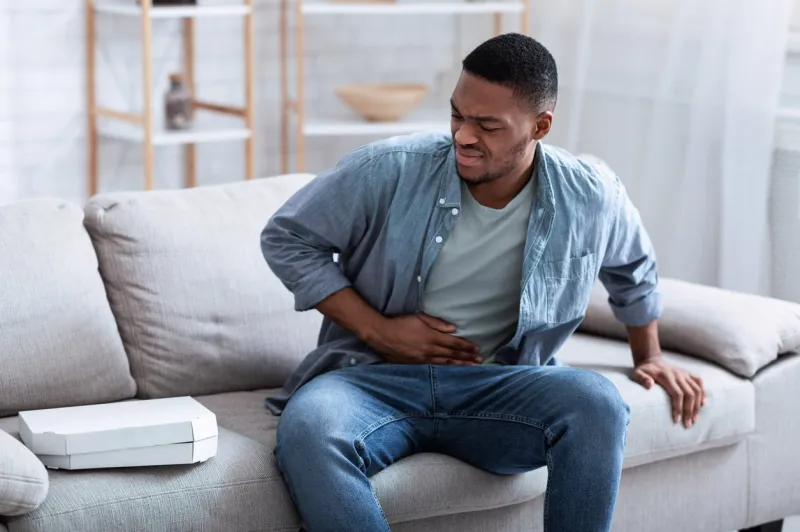 african american guy having stomachache after eating pizza touching aching stomach suffering from pain sitting on sofa at home food poisoning, gastritis, abdominal inflammation and appendicitis