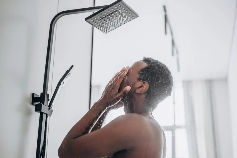 relaxed young african-american man takes shower standing under hot water jets in contemporary unit in bathroom close side view