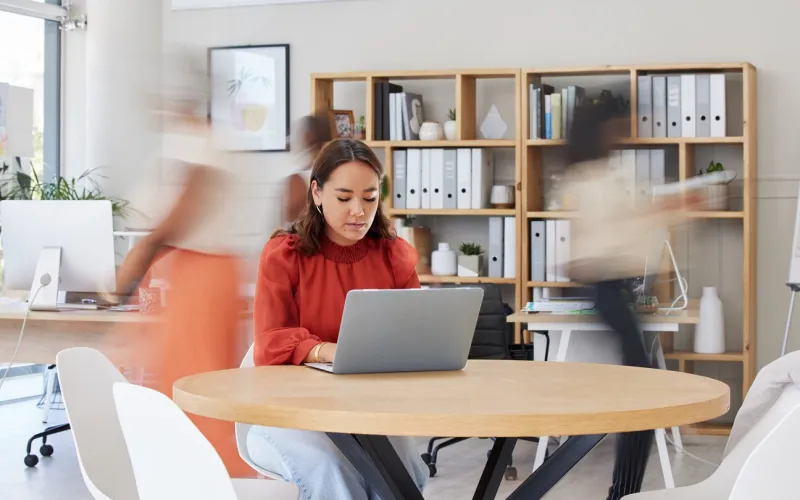 serious asian businesswoman browsing internet on laptop in busy office focused ethnic professional working while colleagues rush in motion blur entrepreneur and creative designer using technology