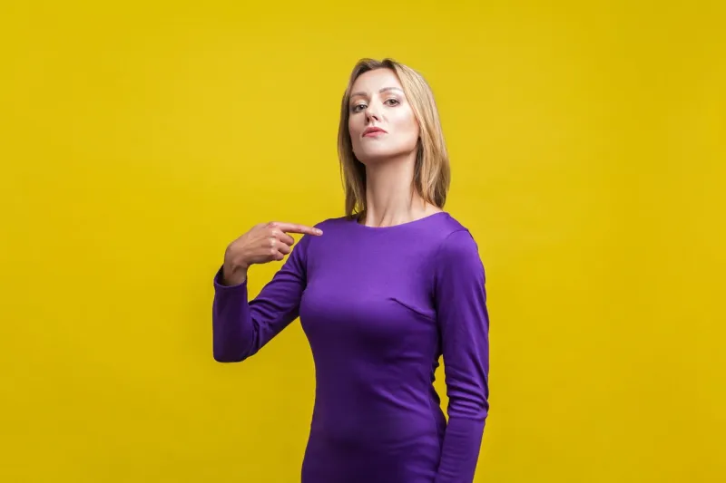 this is me portrait of arrogant businesswoman in tight elegant purple dress pointing at herself, looking egoistic and haughty, proud of achievement indoor studio shot isolated on yellow background