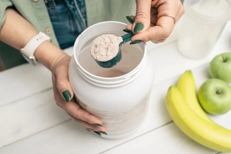 woman in jeans and shirt holding measuring spoon with portion whey protein powder above plastic jar on white wooden table with shaker, banana and apple fruit process of making protein drink