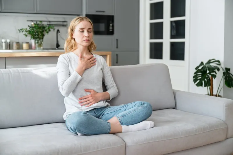mindful young woman with hands on chest and stomach breathing out with closed eyes, calming down at home in stressful situation managing stress, reaching inner balance