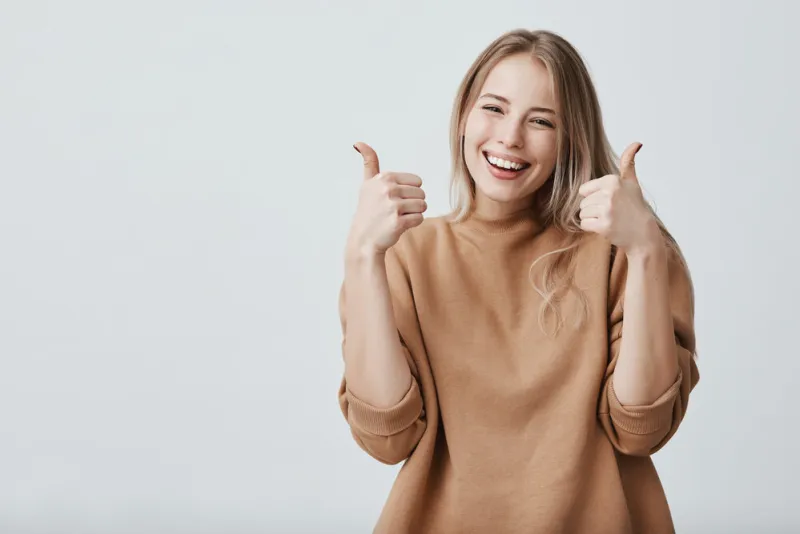 portrait of fair-haired beautiful female student or customer with broad smile, looking at the camera with happy expression, showing thumbs-up with both hands, achieving study goals body language