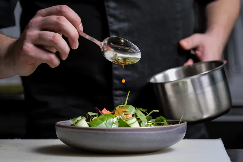 close-up of the hands of a male chef on a black background pour sauce from the spoon on the salad dish food decoration