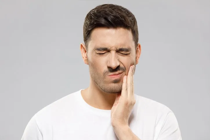 young man in white t-shirt suffering from severe toothache, touching cheek with fingers, eyes closed because of strong pain, isolated on gray background