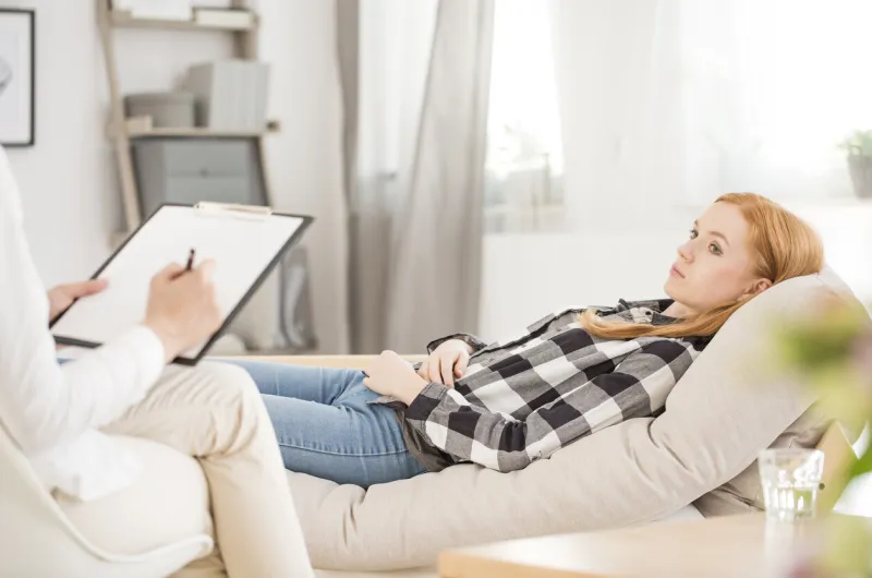 depressed woman lying on sofa during psychotherapy session, psychologist making notes in medical card