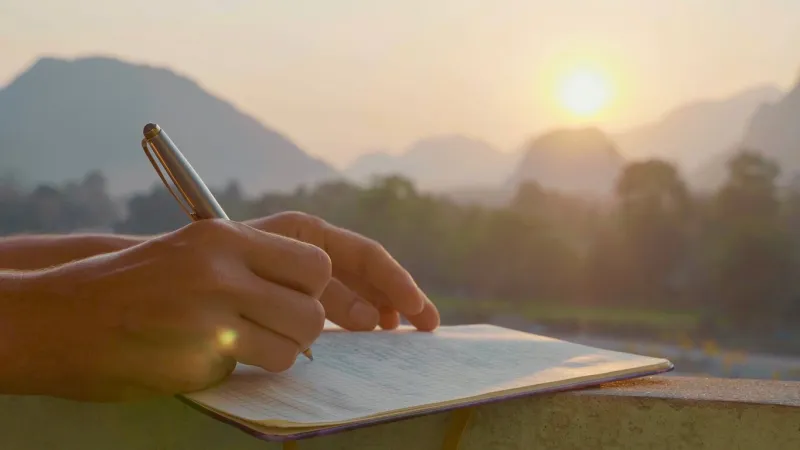 young woman writing travel notes in diary during sunrise with beautiful sun light and mountain landscape on the background, close-up