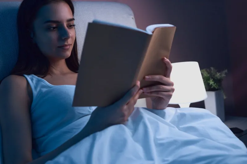 peaceful female lying in bed under soft blanket and reading interesting story in book while spending evening in bedroom before falling asleep