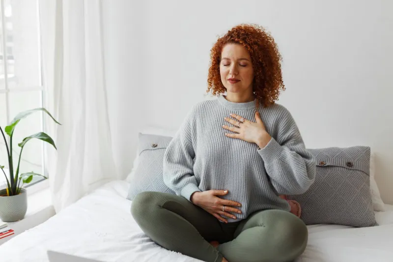 portrait of plus-size curly redhead female in casual clothes doing pranayama technique sitting on bed next to window with closed eyes, putting hands on chest, breathing slowly and deeply