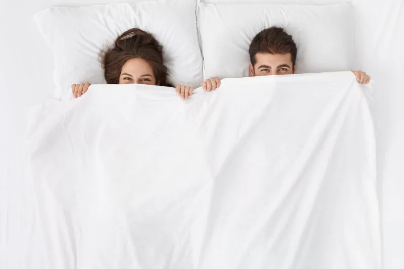 funny married couple lying in bed and hiding under white blanket, looking at camera with eyes full of joy attractive caucasian man and woman having fun in bedroom love and happiness concept