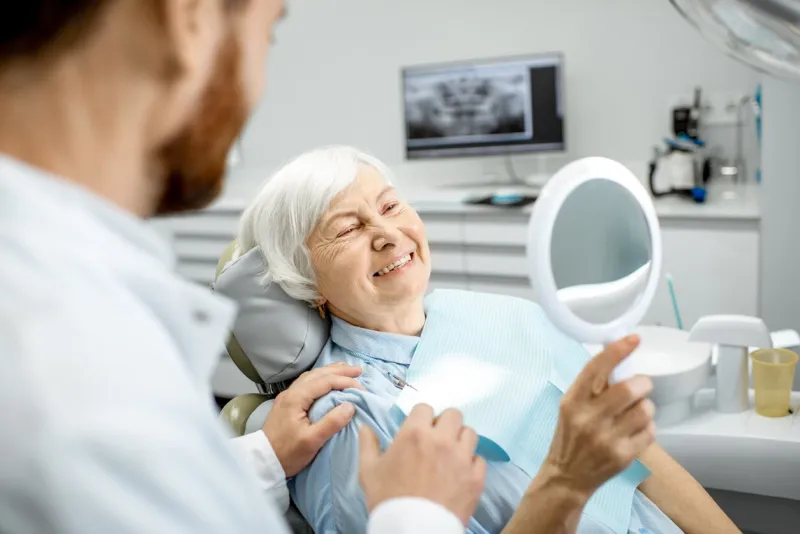 happy elderly woman enjoying her beautiful toothy smile looking to the mirror in the dental office