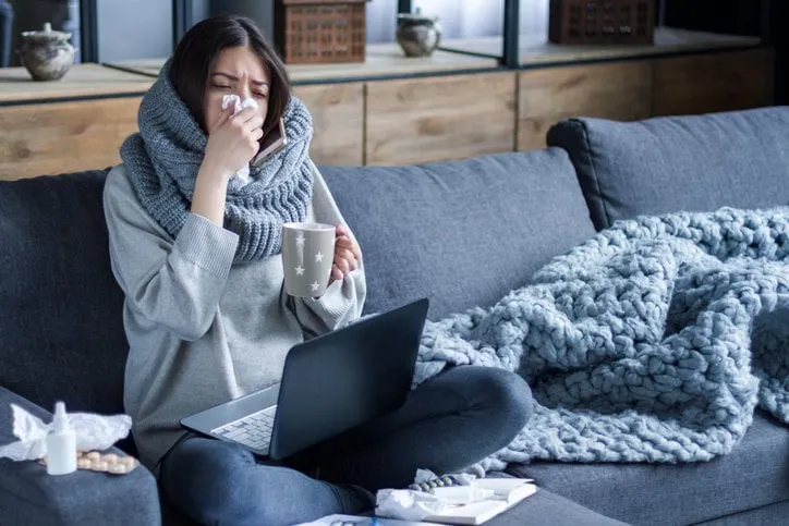 ill brunette girl is sitting at sofa and working remotely on laptop female is blowing out snot, having fever and headache young woman is treated at home, took sick leave winter cold and flu concept