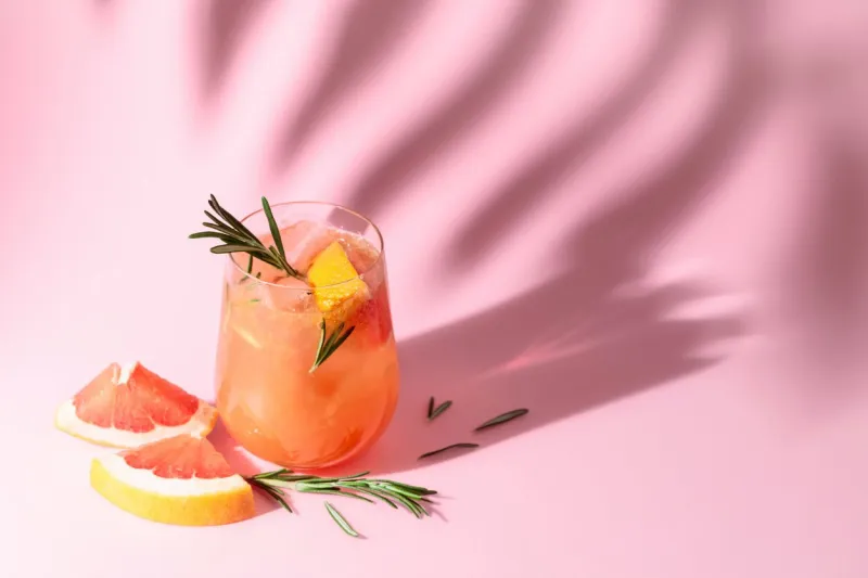 summer cocktail with grapefruit, rosemary, and ice drink on pink background with palm leaf shadow summer, tropical, fresh cocktail concept