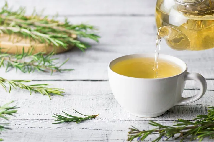 white cup of healthy rosemary tea pouring from teapot with fresh rosemary bunch on white wooden rustic background, winter herbal hot drink concept, salvia rosmarinus