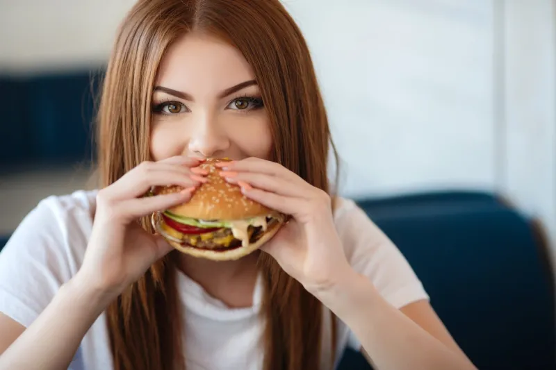 beautiful woman with red long straight hair, bright make-up, brown eyes, red lipstick, long eyelashes, pink nail polish in the hands holding a large hamburger, sitting at a table in a cafe, dinner alone