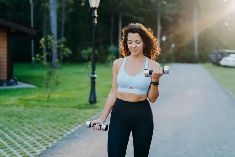 outdoor shot of motivated brunette young woman wears cropped bra and leggings, has slim perfect figure, raises dumbbells and trains biceps, has workout in open air fitness exercising concept