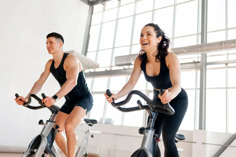 beautiful athletic couple of cyclists train in the fitness room on static bicycle simulator, man trainer and fitness woman go in for sports together on simulators in the morning in bright room