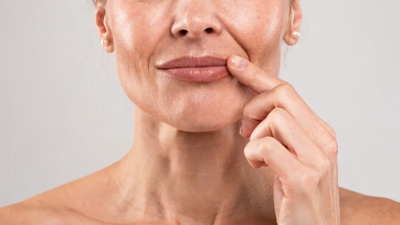 closeup shot of middle aged woman with wrinkles on face touching lips with finger, unrecognizable mature female with bare shoulders posing over light grey studio background, free space