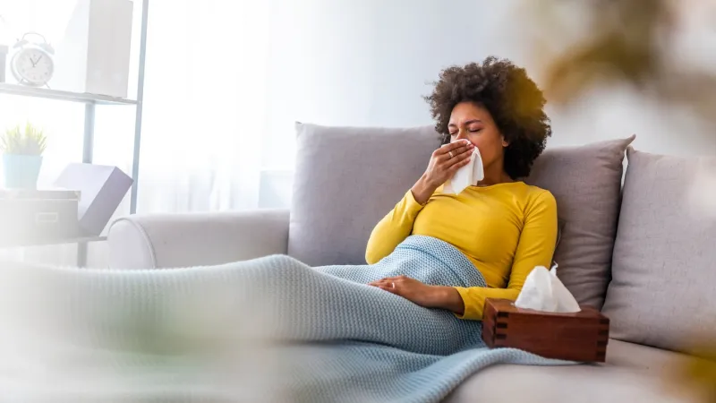 sick young woman sitting on sofa blowing her nose at home in the sitting room photo of sneezing woman in paper tissue picture showing woman sneezing on tissue on couch in the living-room
