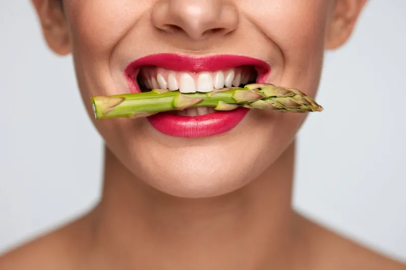 healthy eating closeup of woman mouth with beautiful pink lips makeup holding fresh asparagus between white teeth smiling female face with vegetable in mouth diet food concept high resolution