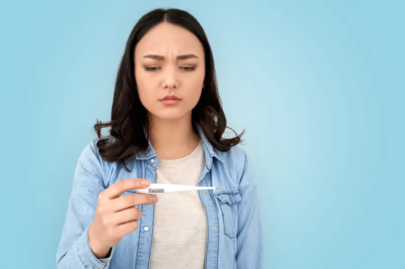 healthcare and treatment concept sick young adult asian woman checking temperature with thermometer, standing isolated on grey background with copy space