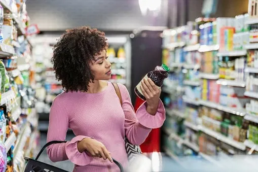 a young african-american woman in her 20s shopping in a grocery store, carrying a shopping basket she is reading the ingredient label on a bottle