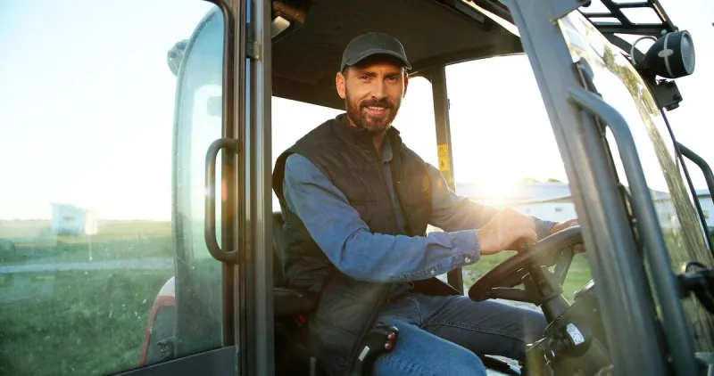 portrait of young caucasian male farmer in cap sitting in tractor with open door and smiling to camera field farming vehicle machine for agriculture handsome smiled man