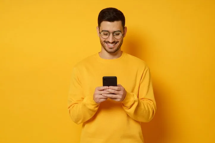 young trendy man in glasses and sweater, feeling happy about conversation in text messages with friends via phone, smiling while looking at screen, isolated on yellow background