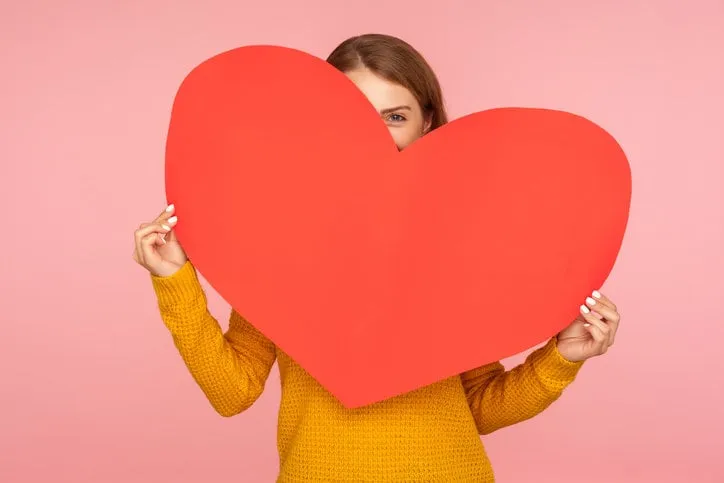 valentine's day portrait of funny girl hiding behind big red heart and looking at camera with playful eyes, holding symbol of love and affection indoor studio shot isolated on pink background