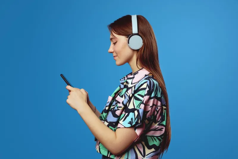 side view of young student girl using mobile phone and looking at screen, wearing headphones while listening to music, isolated on blue background people, lifestyle and emotion concept