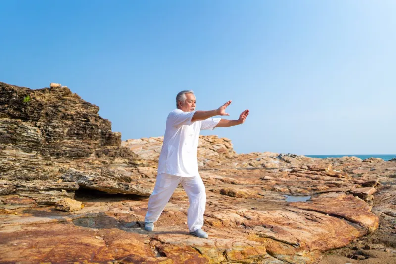 asian senior man practicing meditation with nature on rocky coastal hill retirement elderly people do outdoor relaxing tai chi yoga exercise at summer sunset mental health care motivation concept