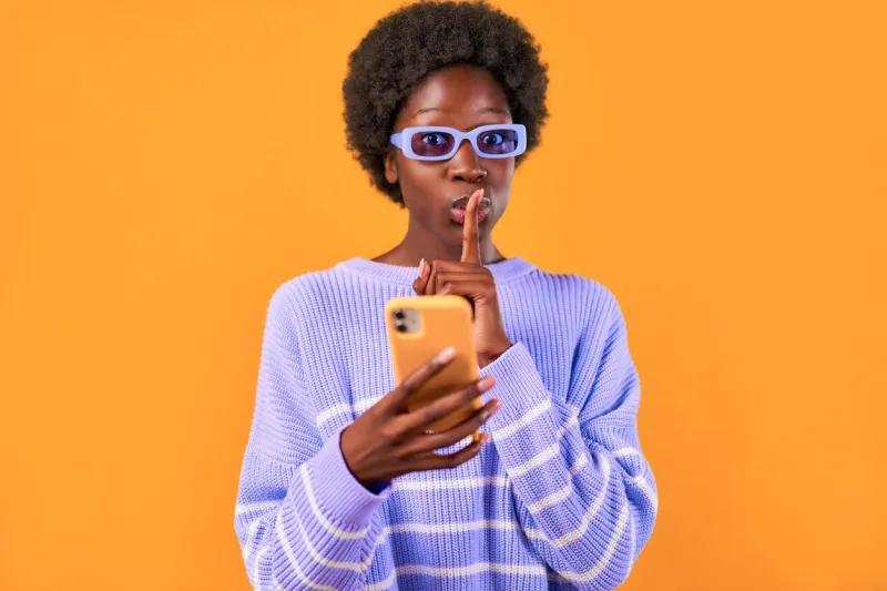 african american young woman with afro hair styling standing in a blue sweater and glasses on a bright orange background using the phone and showing a gesture of quietly placing a finger to her mouth